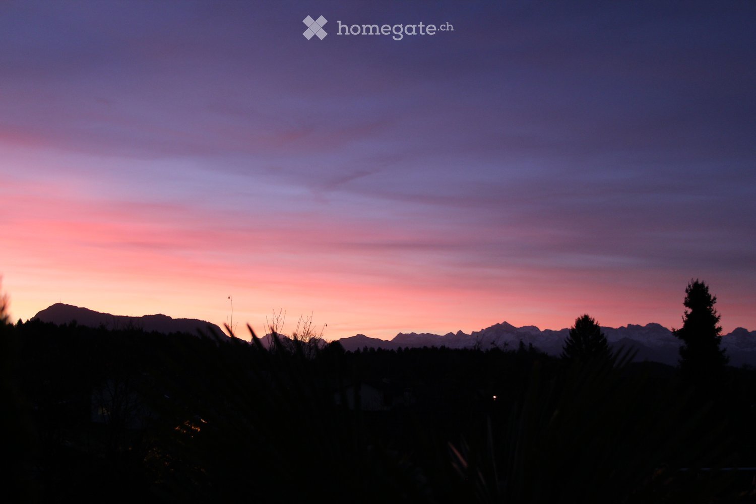 A serene outdoor view at sunset, featuring a mountain range silhouetted against a colorful sky with hues of pink, purple, and orange. The foreground includes dark silhouettes of trees and shrubs.