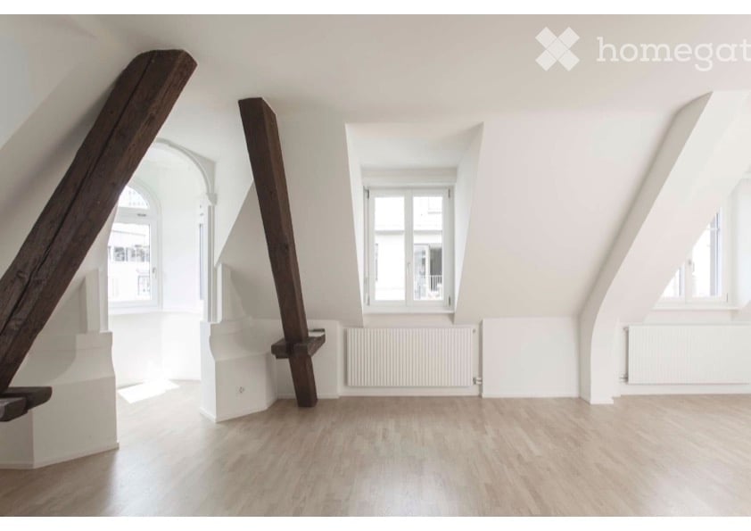 Bright, empty attic room with wooden flooring, white walls, exposed wooden beams, and large windows allowing natural light.