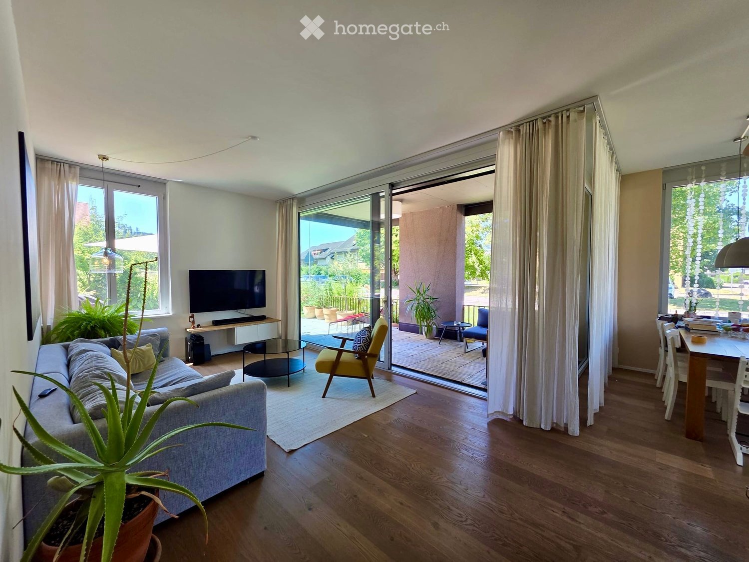 Bright living room with wooden flooring, large windows, and sliding glass doors leading to a balcony. Features a grey sofa, coffee table, dining table, and potted plants.