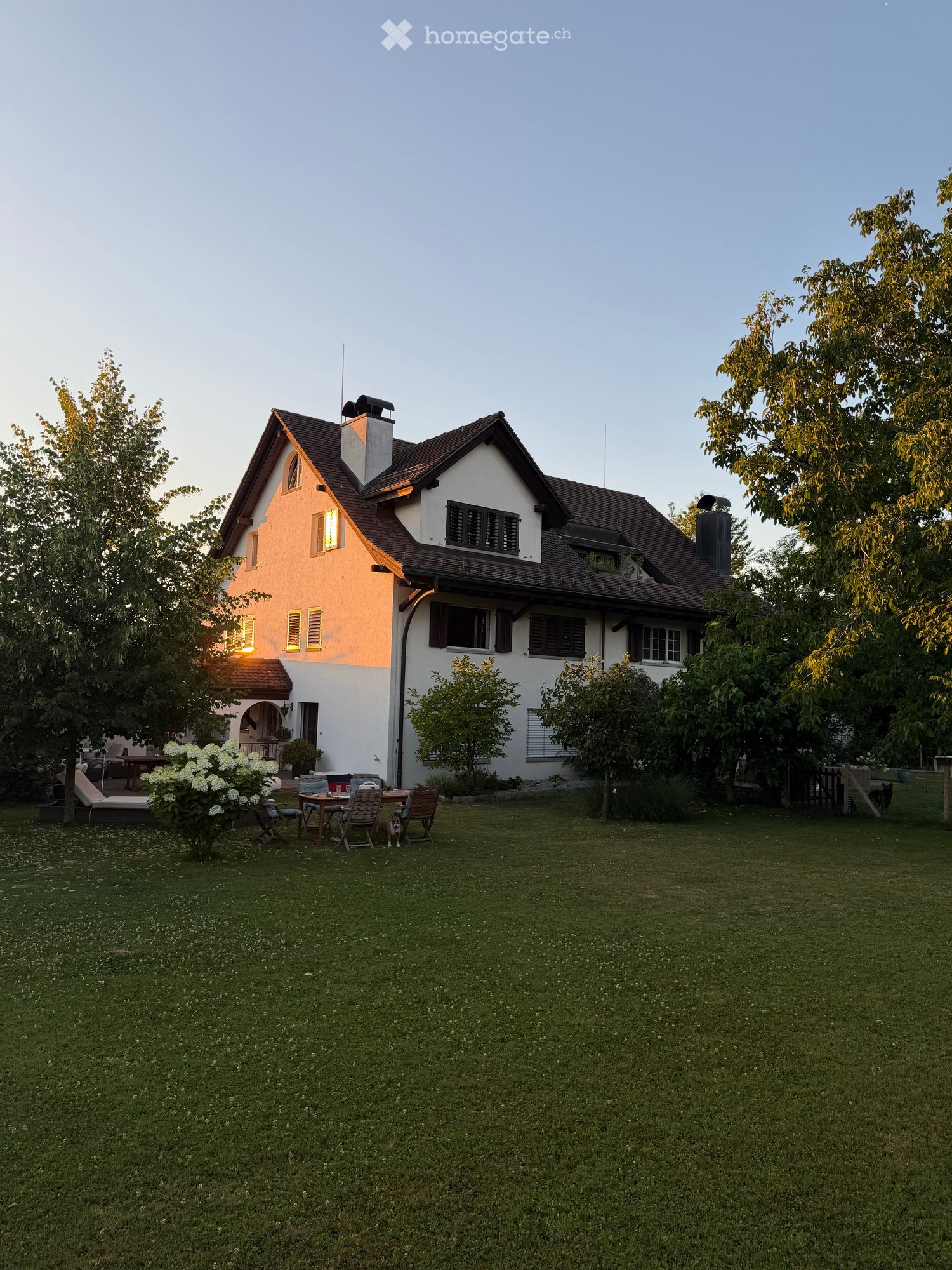 Two-story house with a well-maintained lawn, outdoor seating area, and garden. The house features a mix of white and beige exterior walls, multiple windows, and a chimney. The surrounding area includes trees and shrubs.