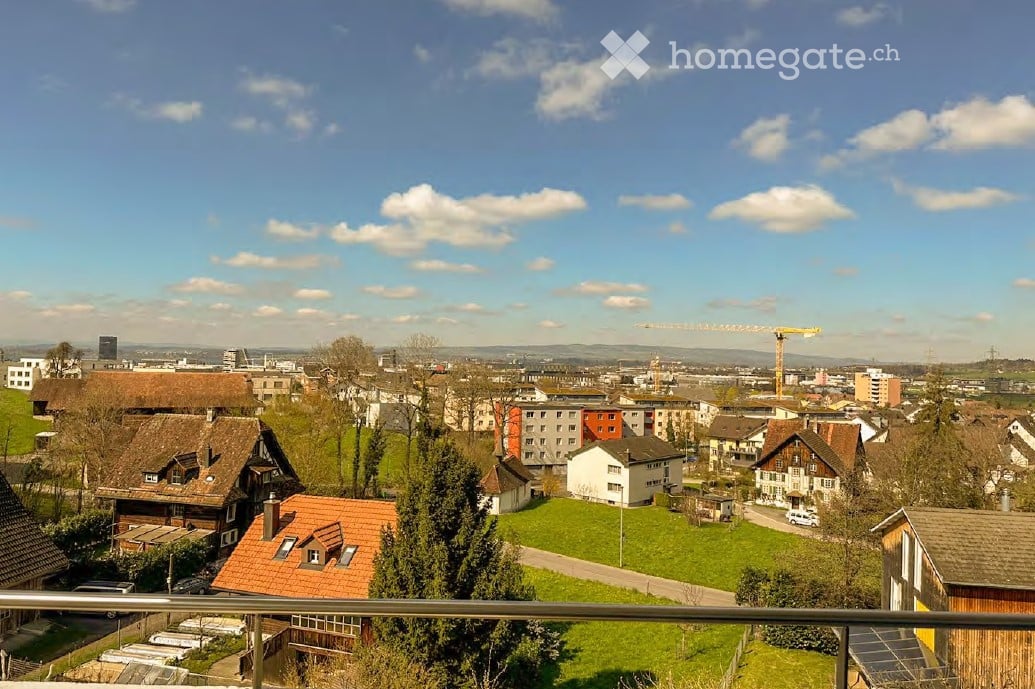 Balcony view overlooking a suburban neighborhood with houses, trees, and a distant cityscape under a partly cloudy sky.