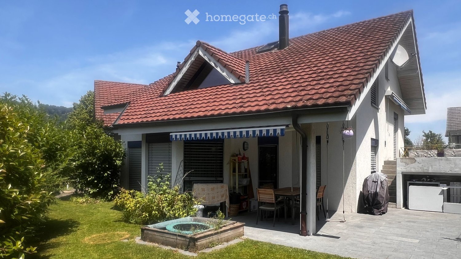 Two-story house with red-tiled roof, white exterior, covered patio with outdoor furniture, small swimming pool, and garden area.