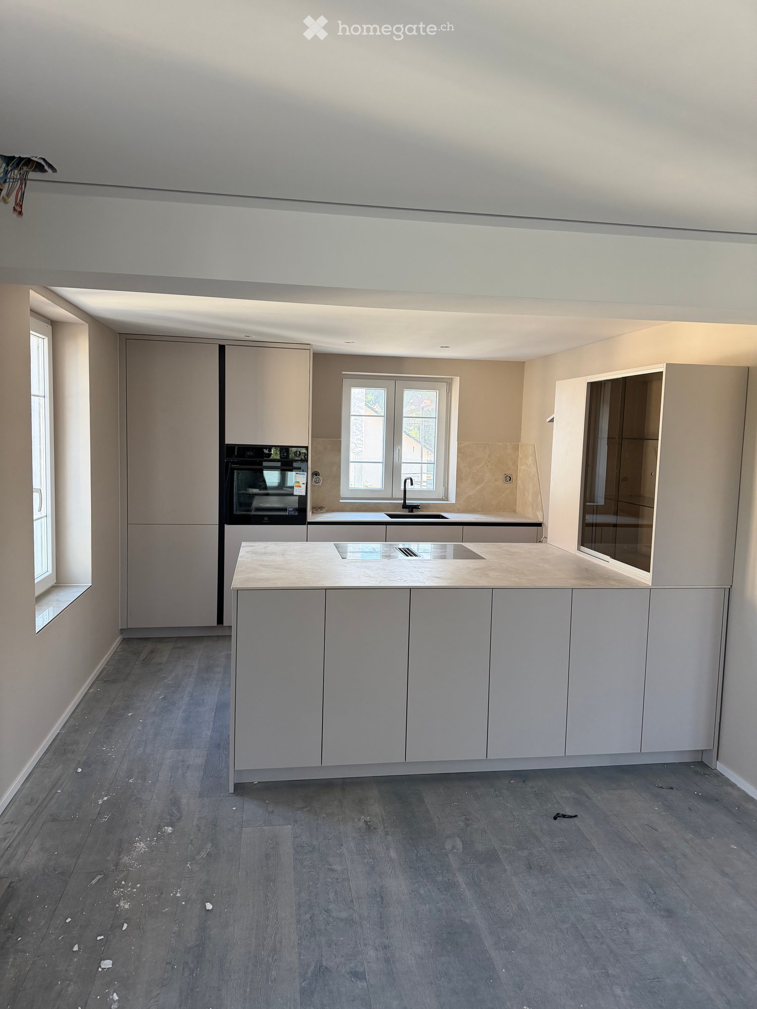 Empty kitchen with light grey cabinets, a central island, and a built-in oven. The floor is made of dark grey wood, and there are windows on two walls.