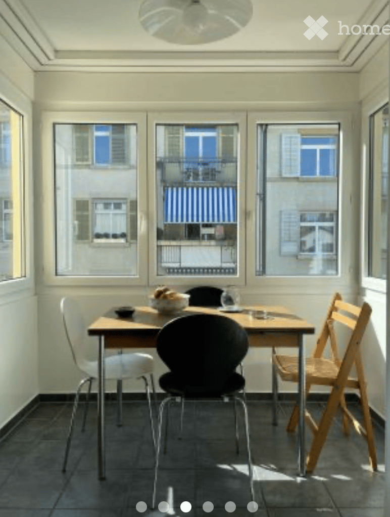 Bright, airy room with large windows offering a view, featuring a wooden dining table with chairs and a bowl of food.