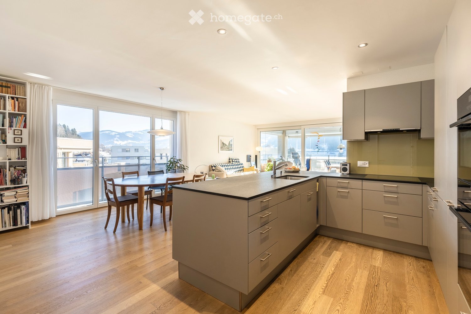 Modern kitchen with a central island, cabinets, and a dining area. Large windows offer a scenic mountain view. Includes a table, chairs, bookshelf, and a small living area with a sofa and plant.