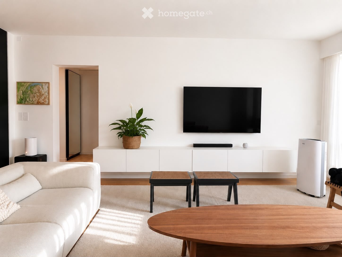 Modern living room with white walls, wooden flooring, a white sofa, a wooden coffee table, two small stools, a potted plant, a mounted TV, and a white cabinet.