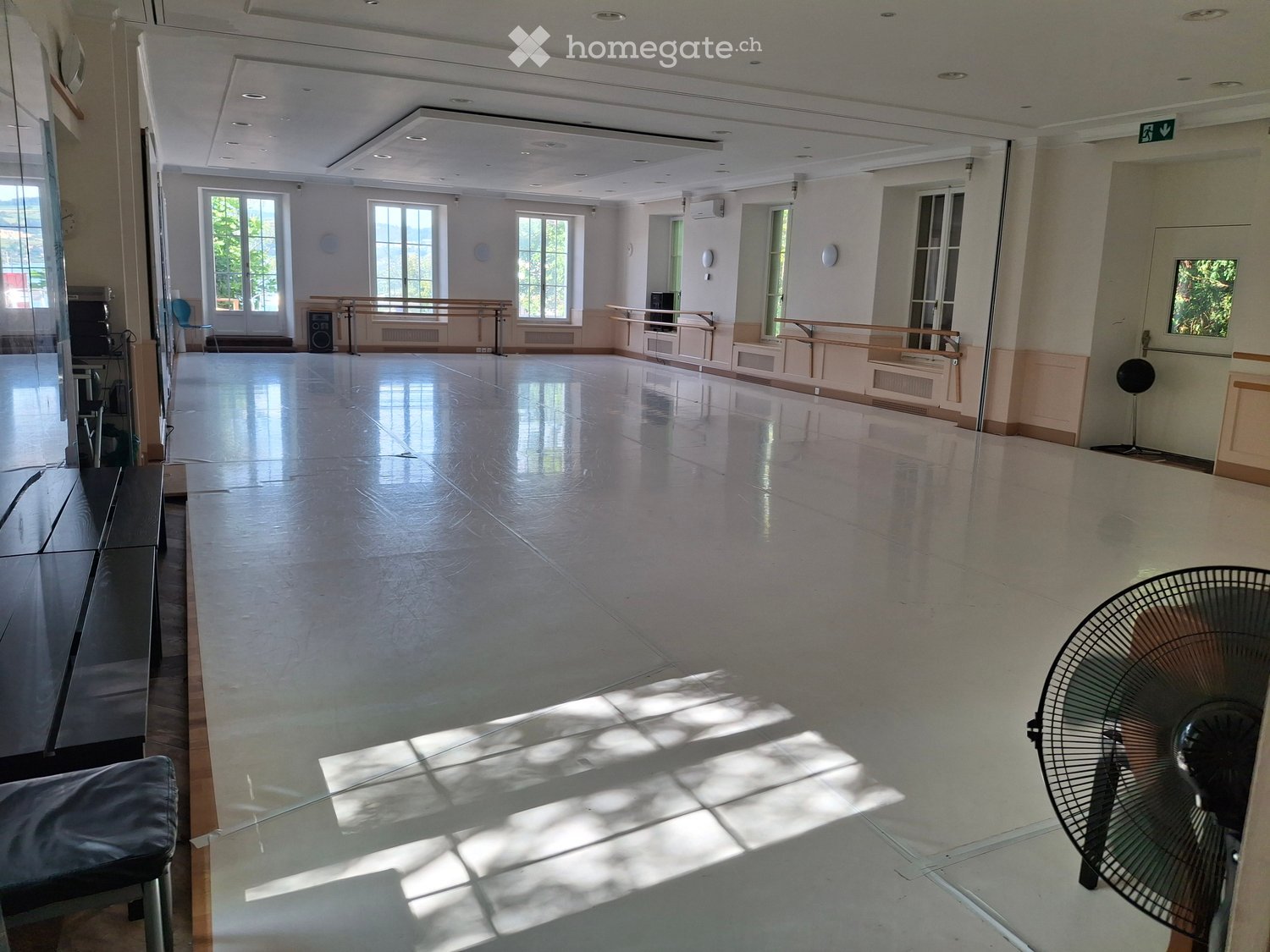 Large, empty room with a polished concrete floor, large windows, and a glass wall. The space is well-lit with natural light and has a fan in the corner.