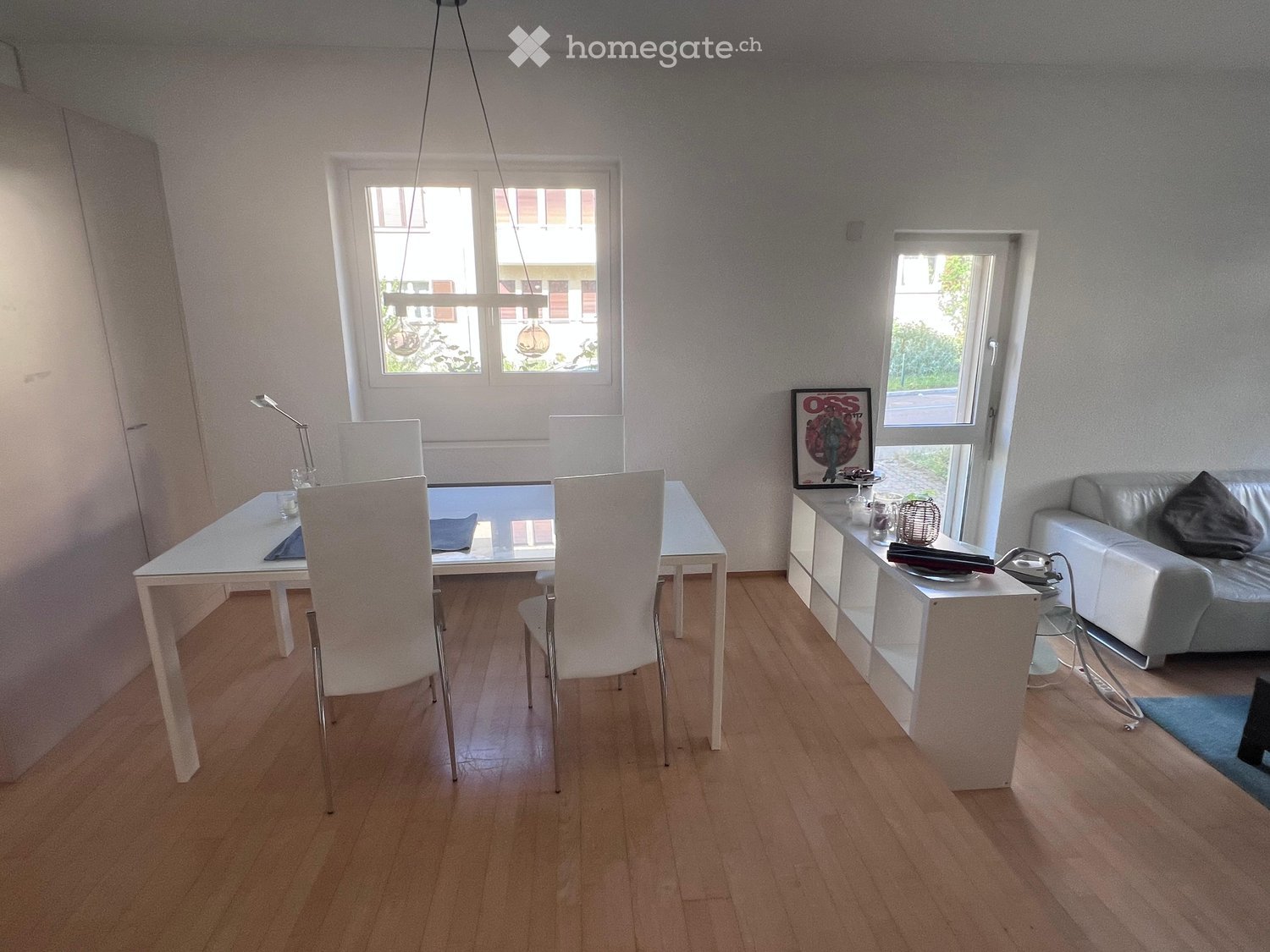 Modern open-plan living and dining area with light wooden flooring, white dining table with chairs, grey sofa, and a small side table with a lamp and picture frame.