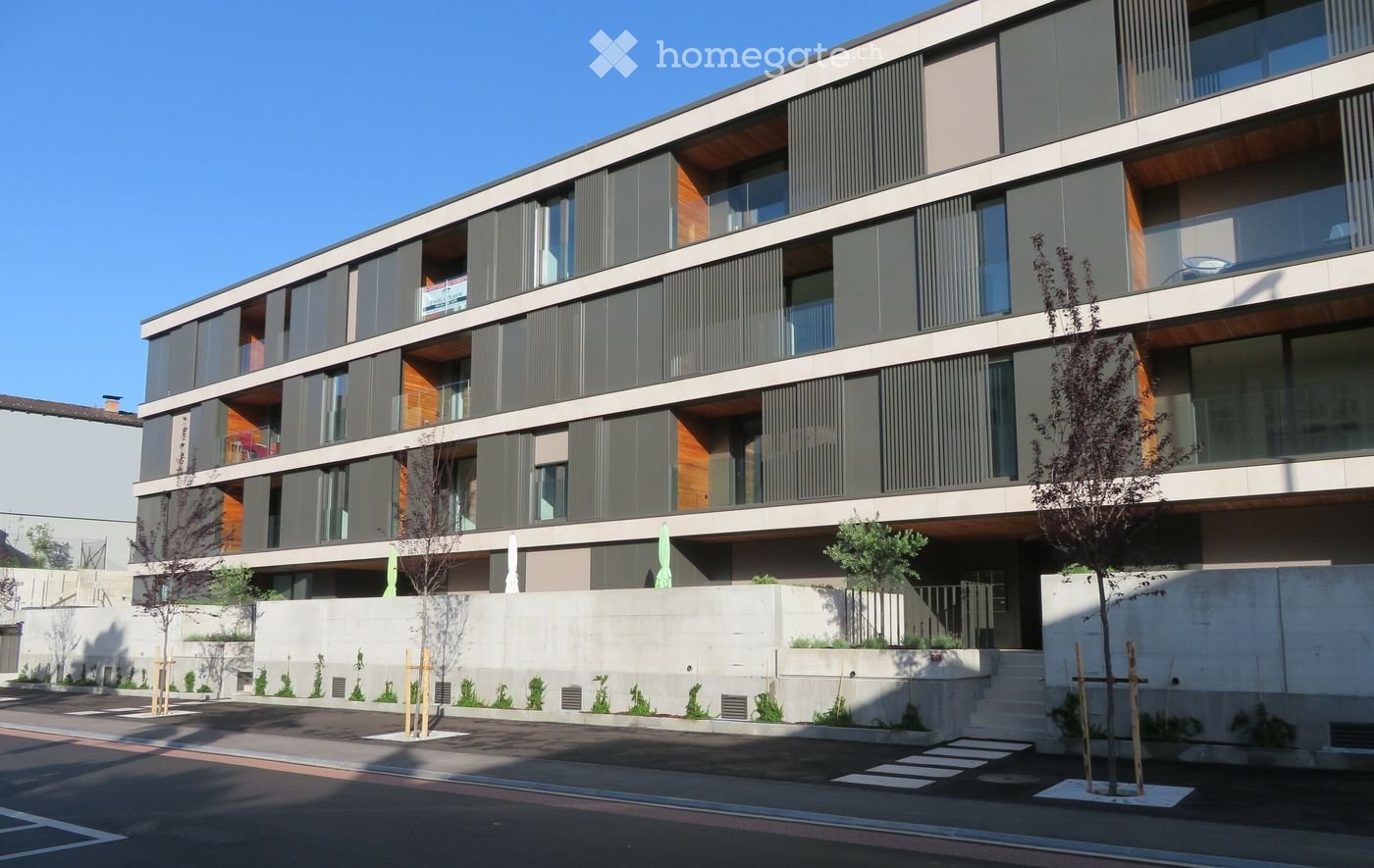 Modern multi-story residential building with large windows, balconies, and contemporary design. Features include a raised ground floor, greenery, and a clean, minimalist aesthetic.