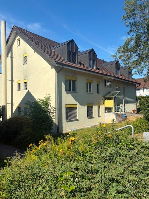 Two-story residential building with light yellow walls, multiple windows with yellow shutters, and a red-tiled roof. The building is surrounded by greenery and has a small garden area in front.