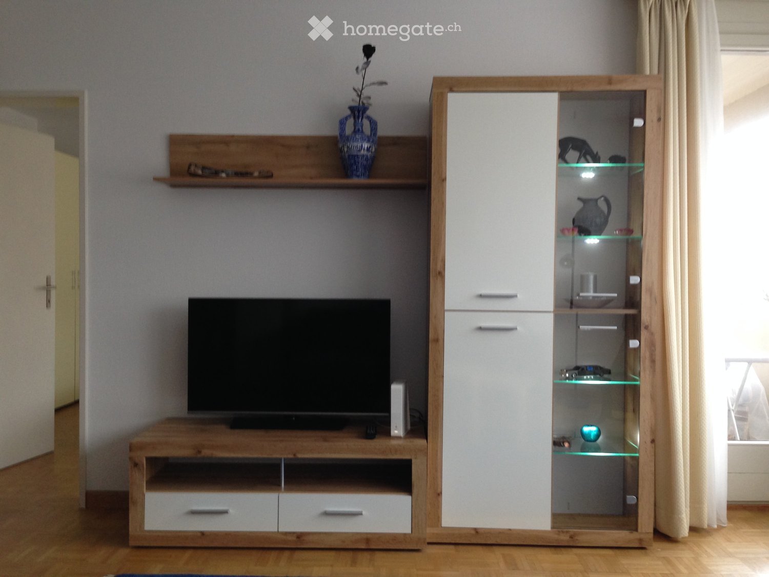 Modern living room with a wooden TV stand, flat-screen TV, white cabinet with glass doors, and decorative vase on a shelf.