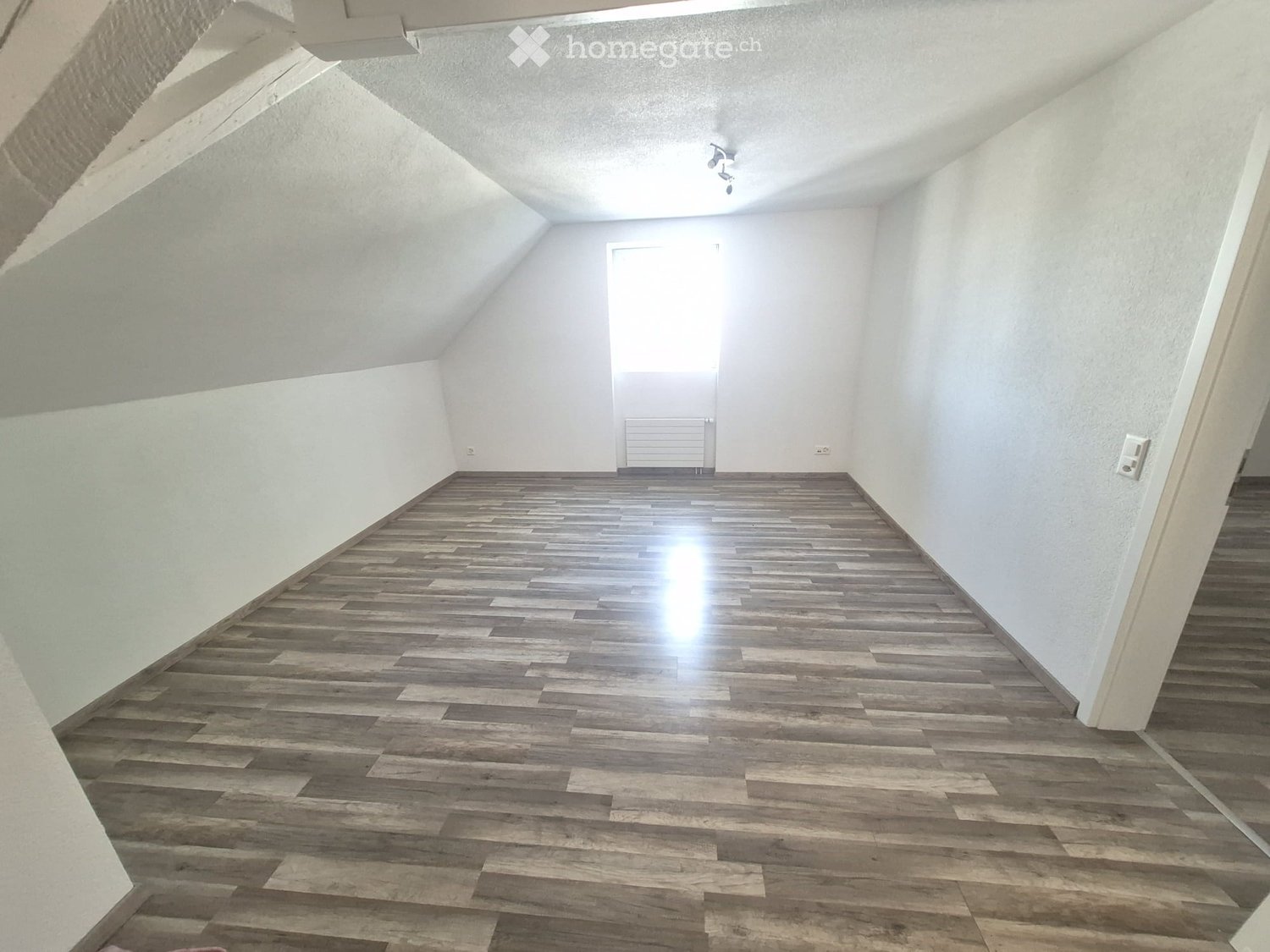 Empty room with wooden flooring, white walls, and a door leading to another space.