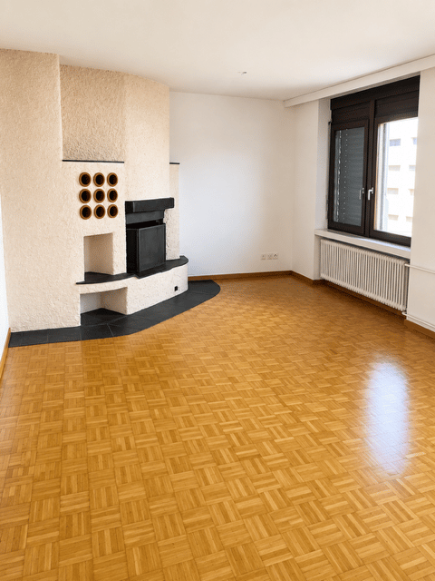 Empty room with wooden parquet flooring, white walls, a fireplace with a black insert, and a large window with blinds.
