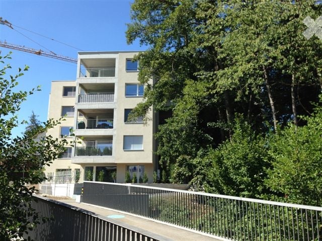 Modern multi-story apartment building with balconies, surrounded by lush green trees.