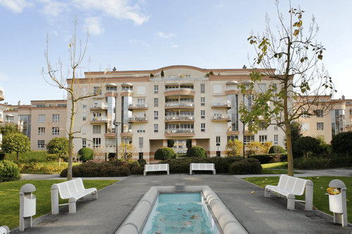 Courtyard area with a small fountain, several white benches, trees, and a multi-story residential building in the background.
