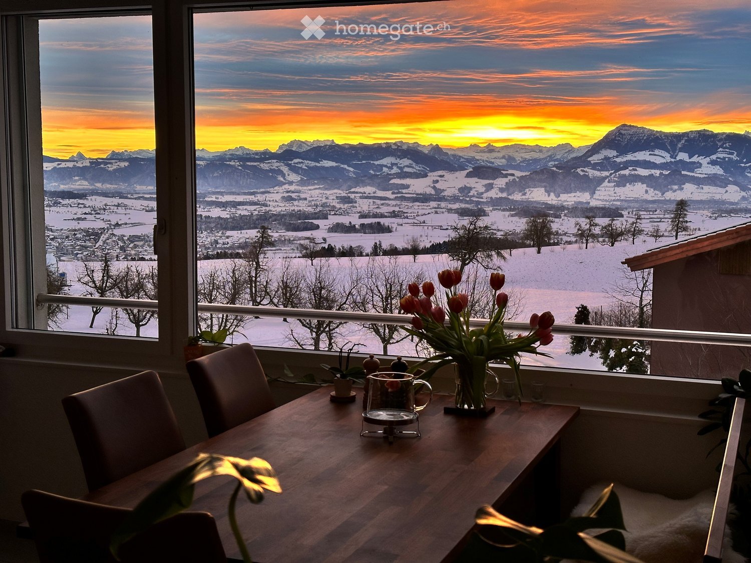 Bright room with large window showcasing a scenic mountain view at sunset, featuring a wooden table with chairs, a vase of tulips, and a cup.