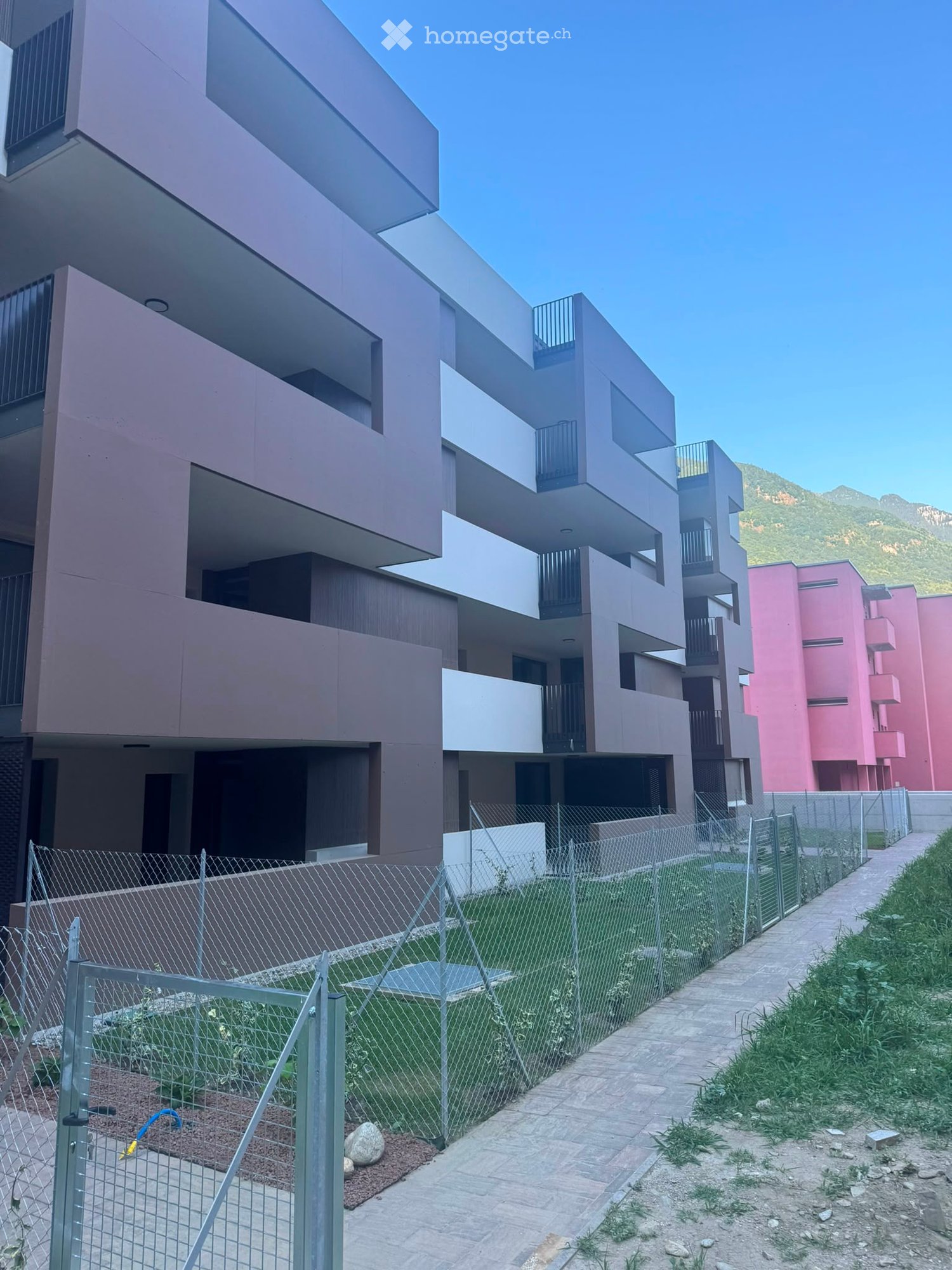 Modern multi-story residential building with balconies, surrounded by a fenced area and pathway.
