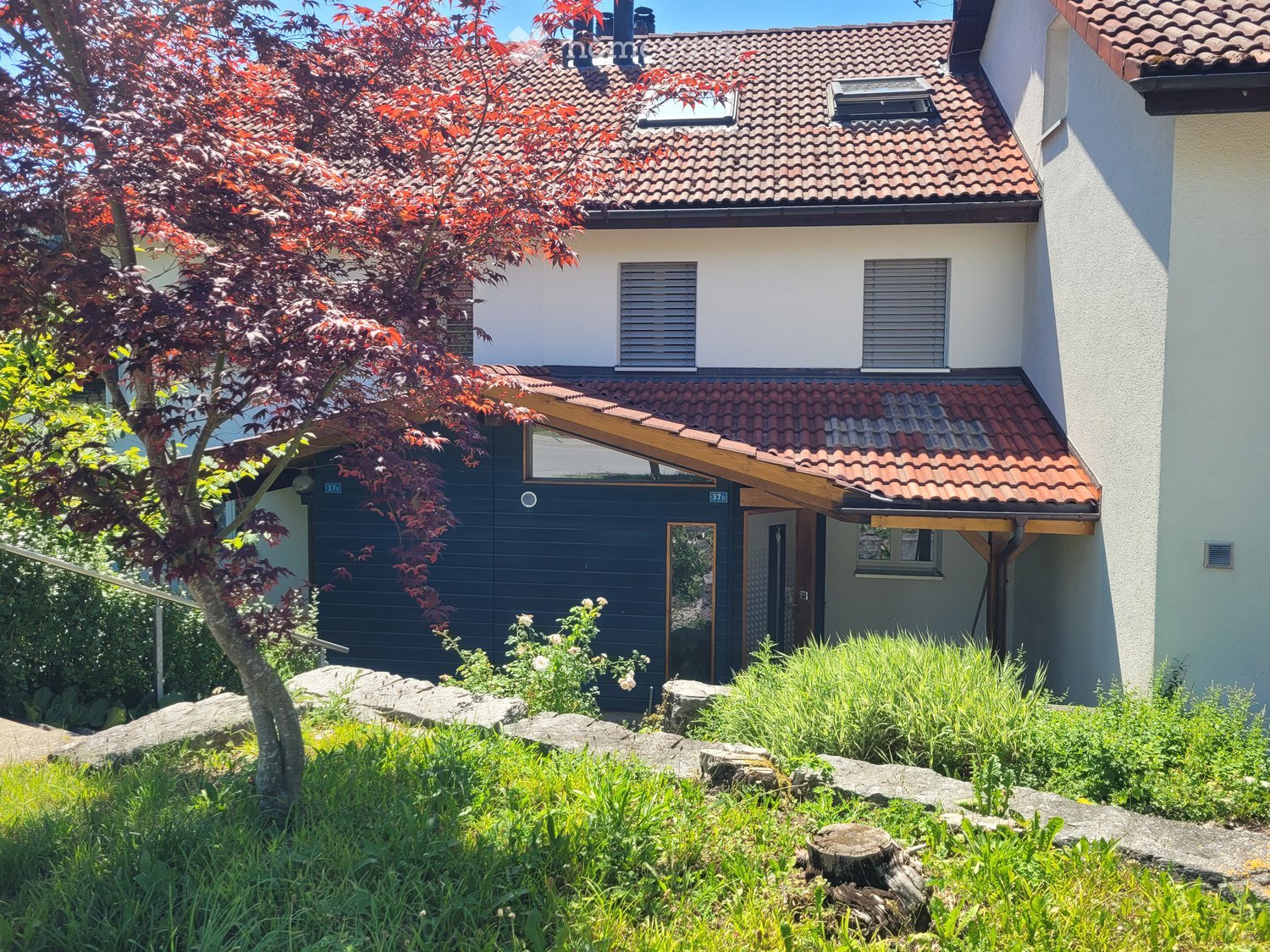 Two-story house with red-tiled roof, white walls, and a dark blue garage. The house features a small porch with a wooden roof and a tree with red leaves in the front yard.