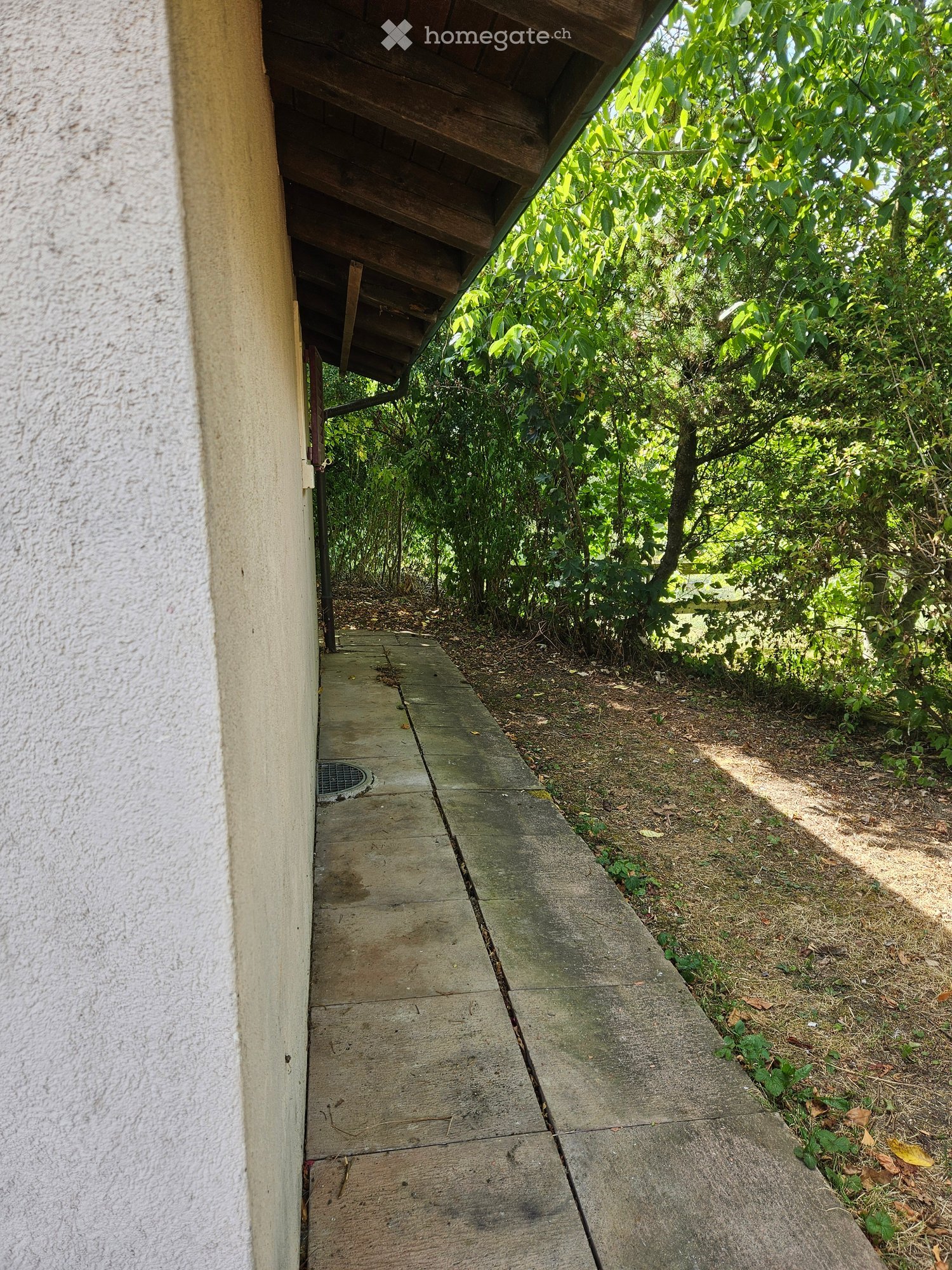 A narrow concrete pathway leading from a covered entrance area into a lush, green garden. The pathway is bordered by grass and small plants, with a wooden roof providing partial shade.