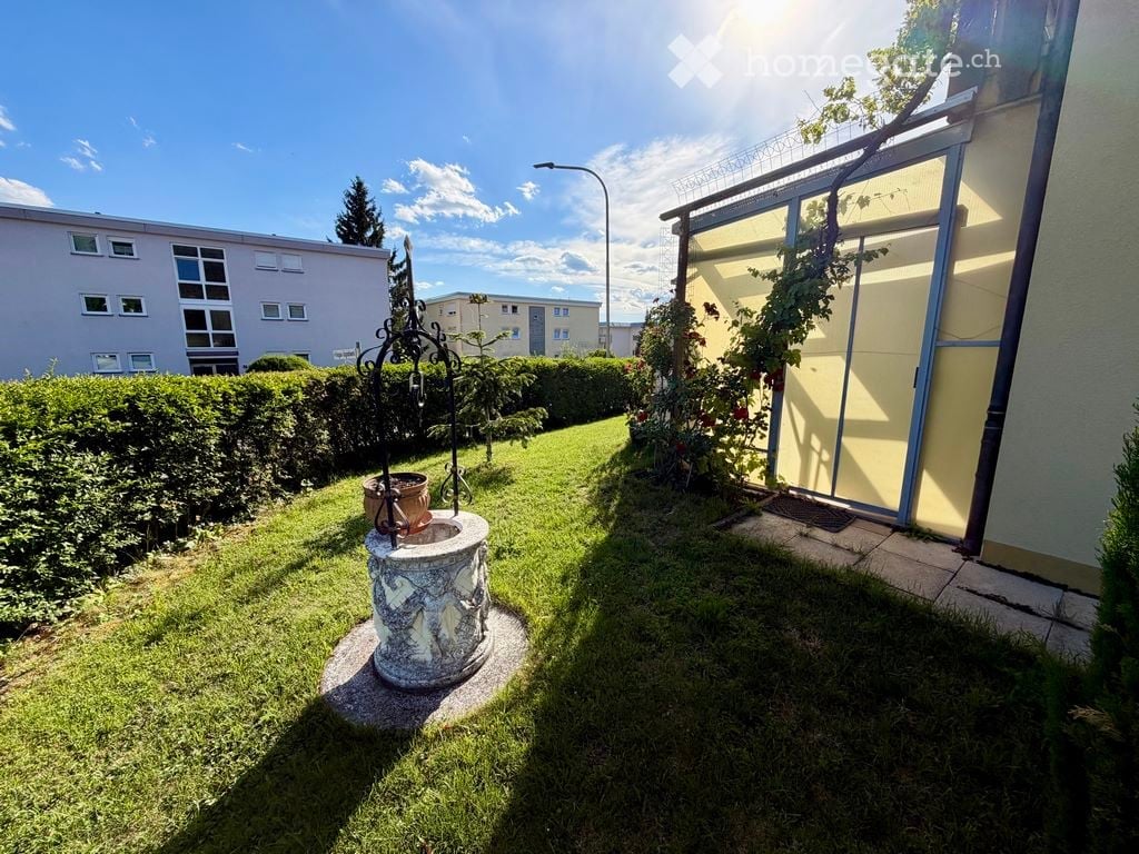 Well-maintained garden with green grass, trimmed hedges, decorative fountain, and a small metal structure with a potted plant. A modern building with large windows and a sliding glass door is visible on the right.