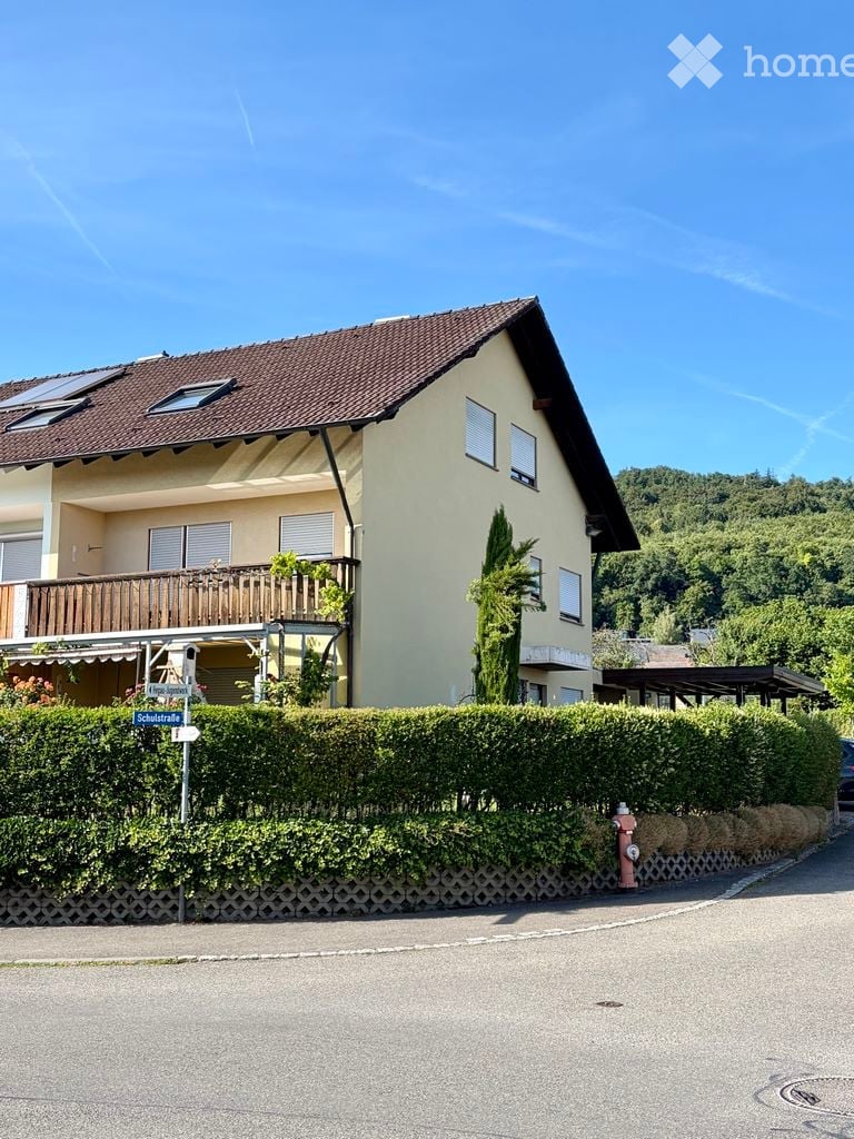 Two-story house with a brown tiled roof, beige walls, and a wooden balcony on the first floor. The house is surrounded by a neatly trimmed hedge and is situated in a residential area with a clear blue sky and green hills in the background.
