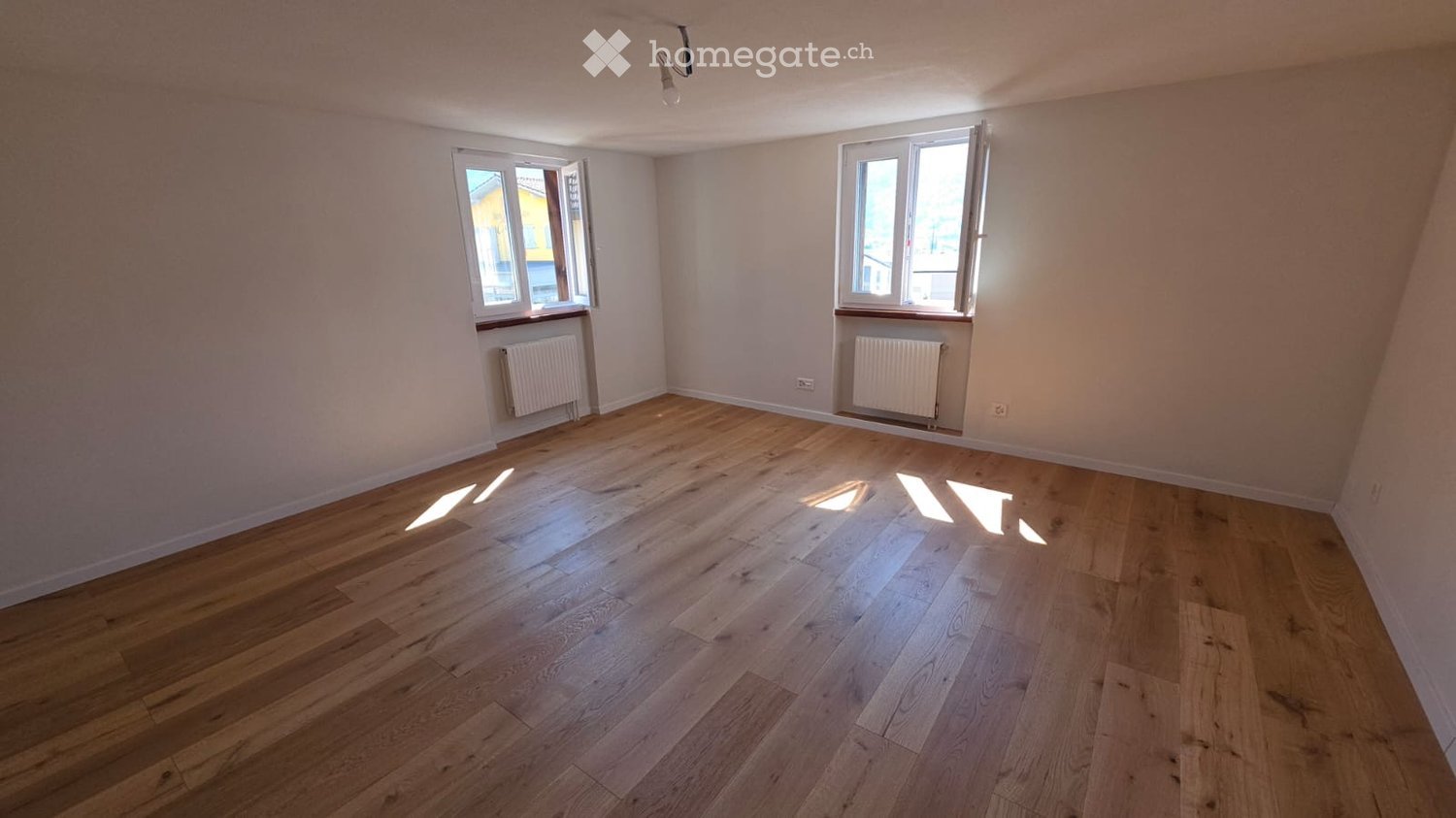 Empty room with wooden flooring, white walls, two windows, and radiators under each window. No furniture or personal items present.