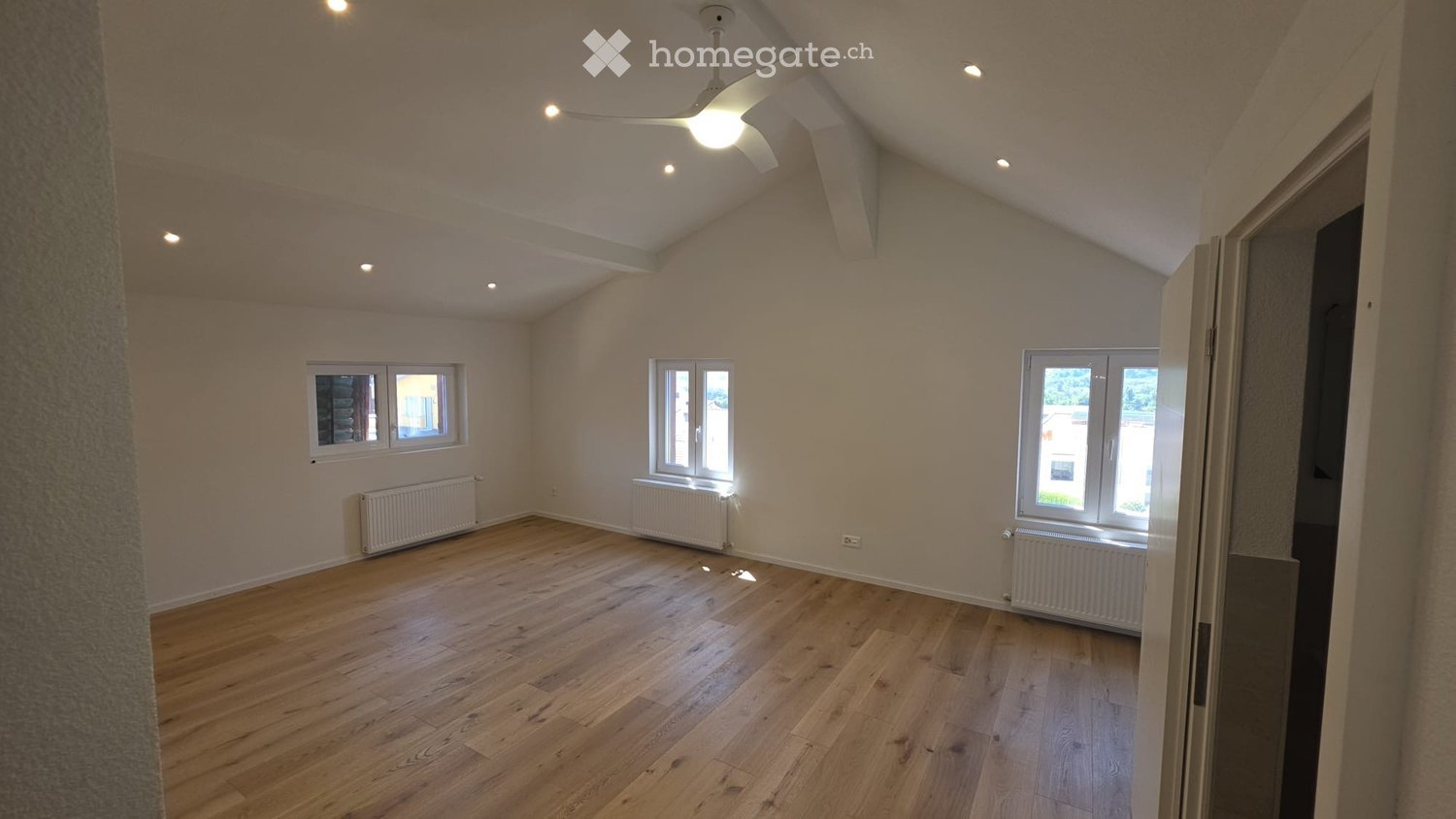 Empty room with wooden flooring, white walls, and multiple windows allowing natural light. Features include a ceiling fan with light, radiators along the walls, and a partially visible adjacent space through an open doorway.