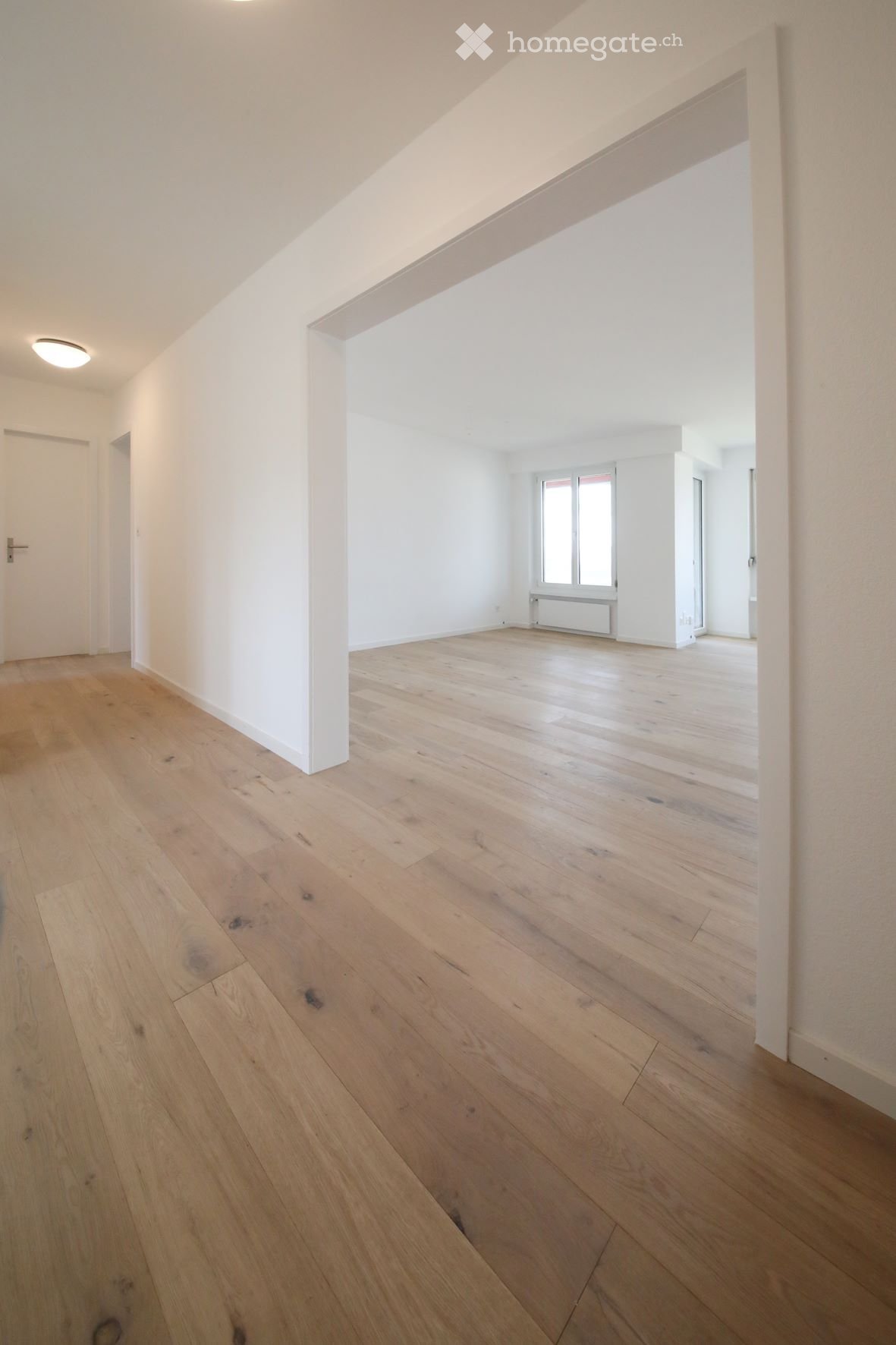 Empty room with light wooden flooring, white walls, and a large window with multiple panes. The room has a simple, modern design with a visible door and a recessed ceiling light.