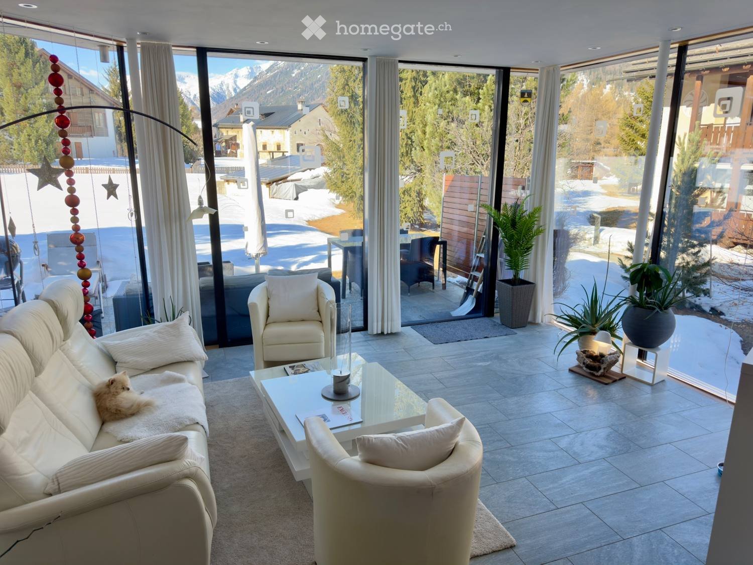 Modern living room with large windows offering a mountain view, featuring a white sofa, coffee table, armchairs, and a small dog resting on the sofa.