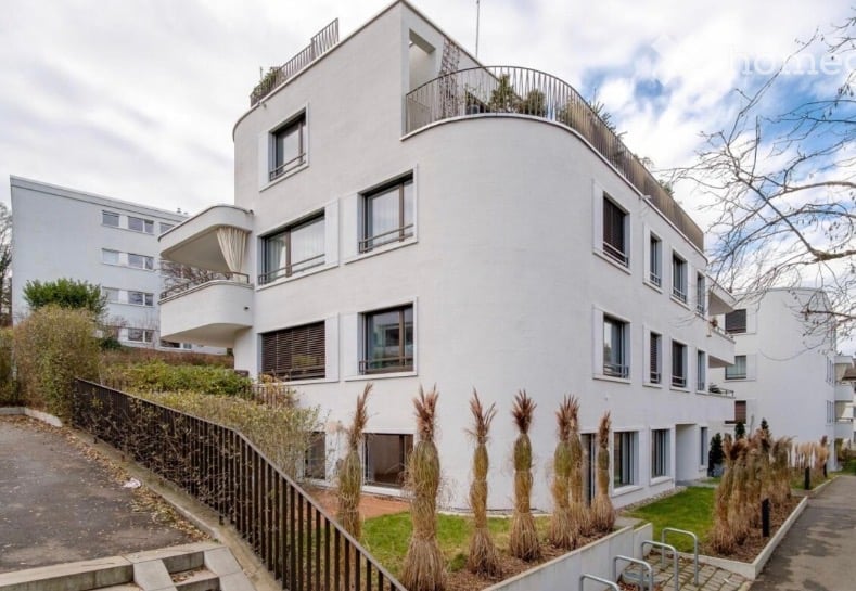 Modern white multi-story building with curved facade, multiple balconies, and a rooftop terrace.