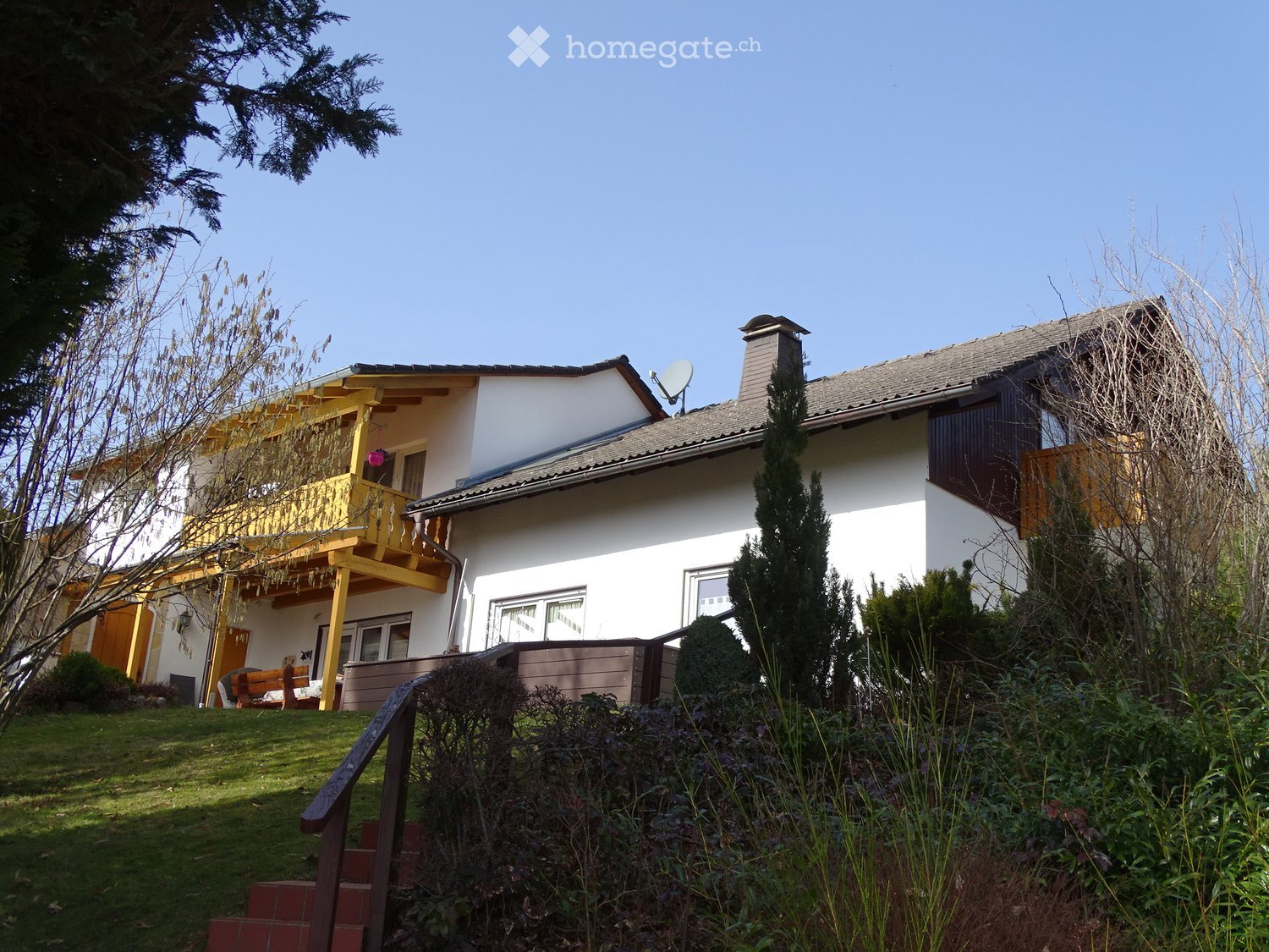 Two-story house with white walls, grey roof, yellow balcony, and surrounding greenery.