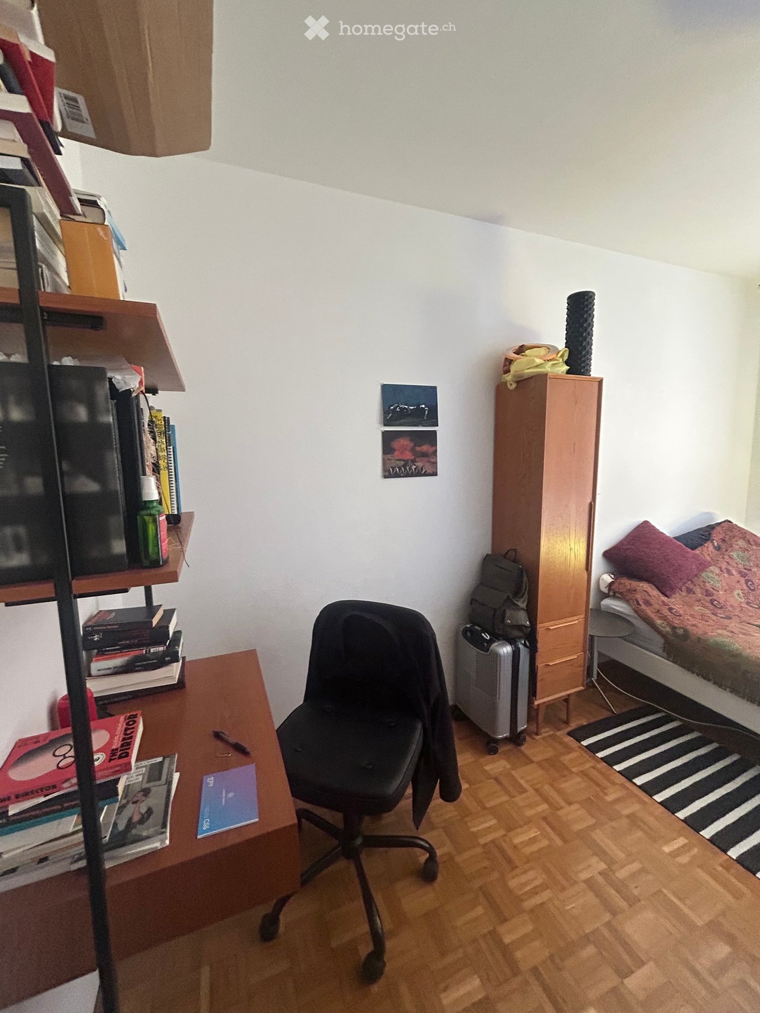 A small, furnished bedroom with a wooden floor, a desk with a chair, a bed with pillows and a blanket, a wardrobe, and a backpack on the floor.