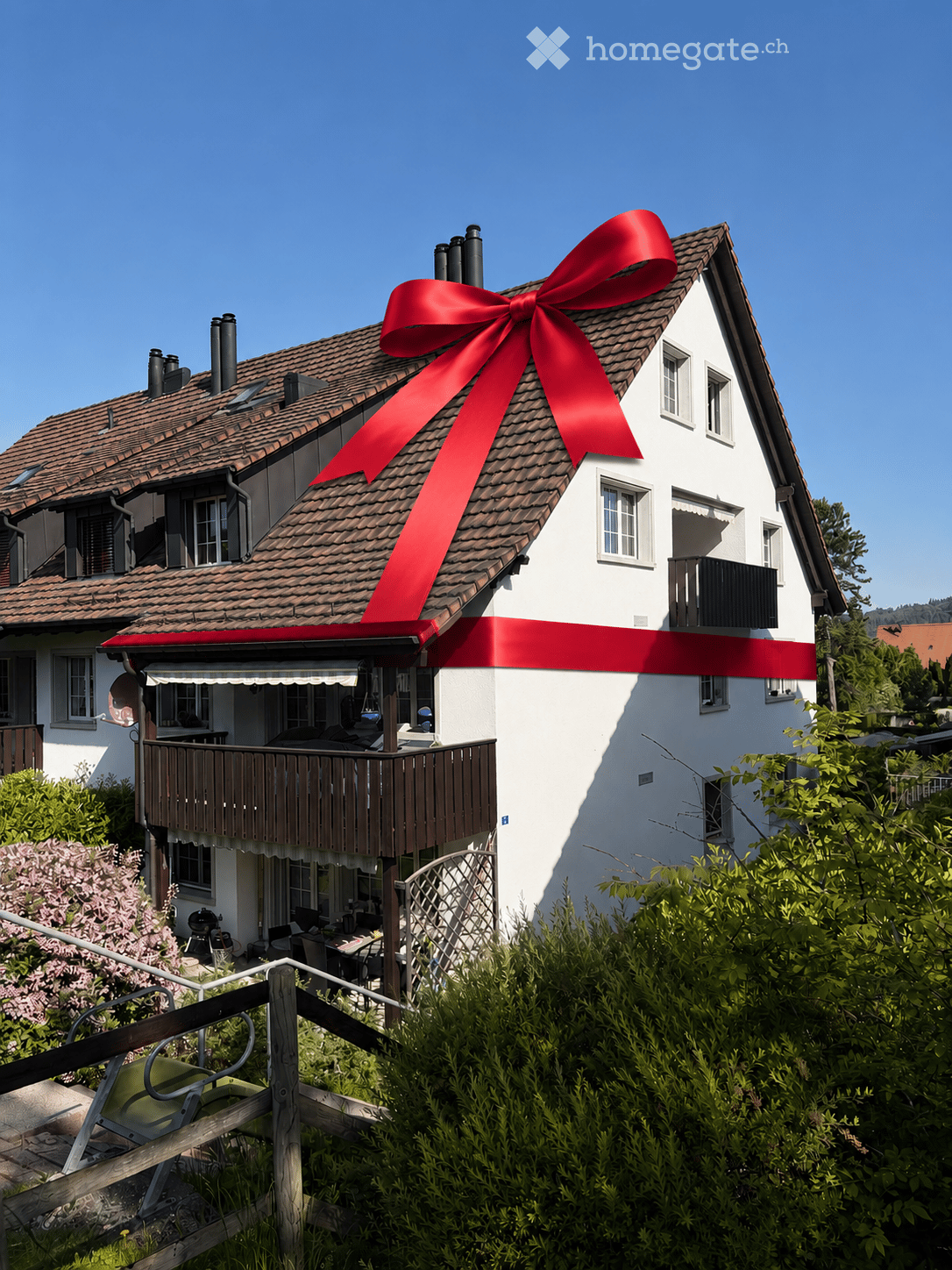 Two-story house with a red roof and white walls, featuring a large red ribbon on the roof, a balcony, and a well-maintained garden.