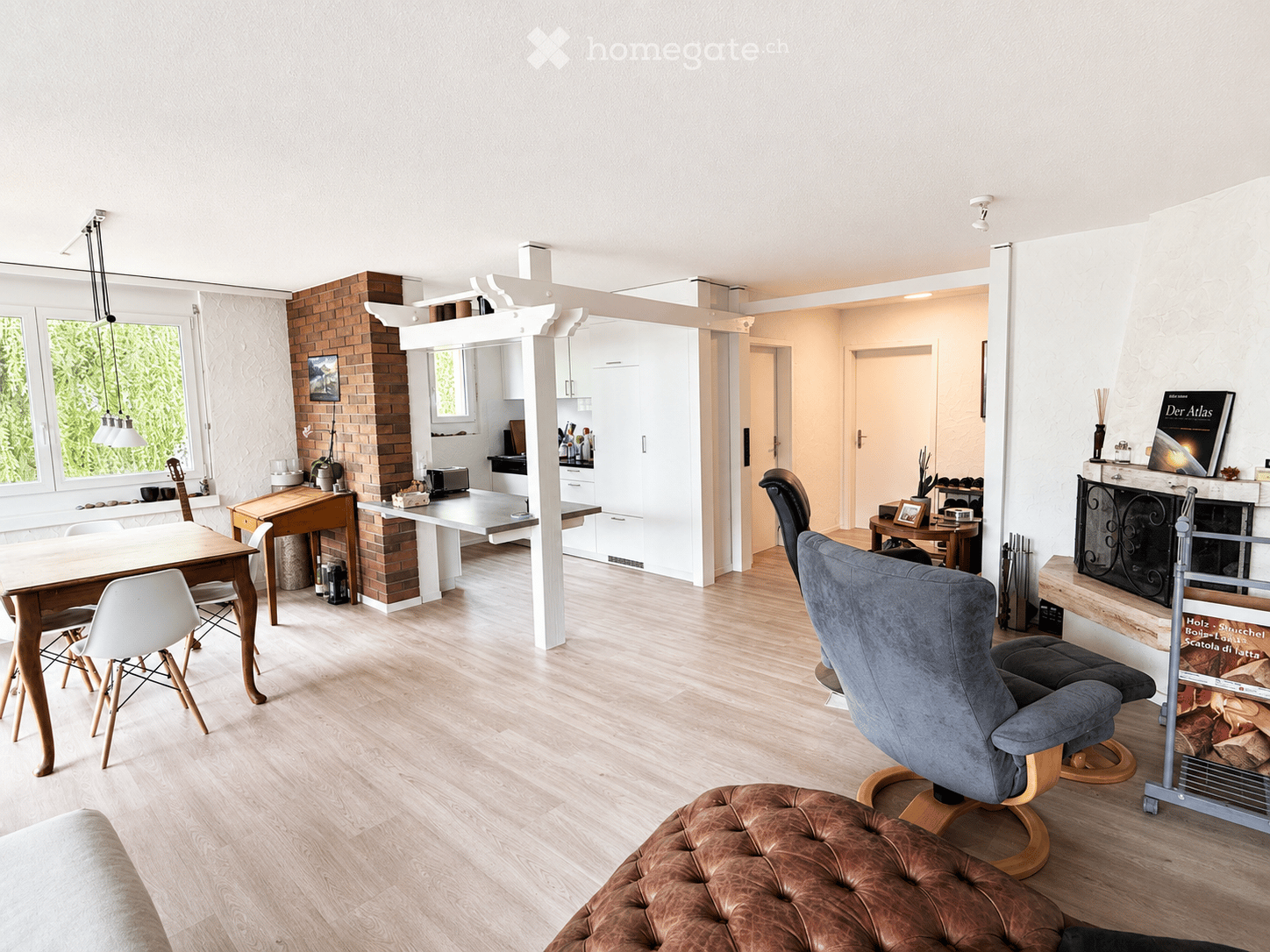 Open-plan living and dining area with light wooden flooring, white walls, and large windows. Features a grey sofa, brown leather ottoman, dining table with chairs, fireplace, and a kitchen area with cabinets and appliances.