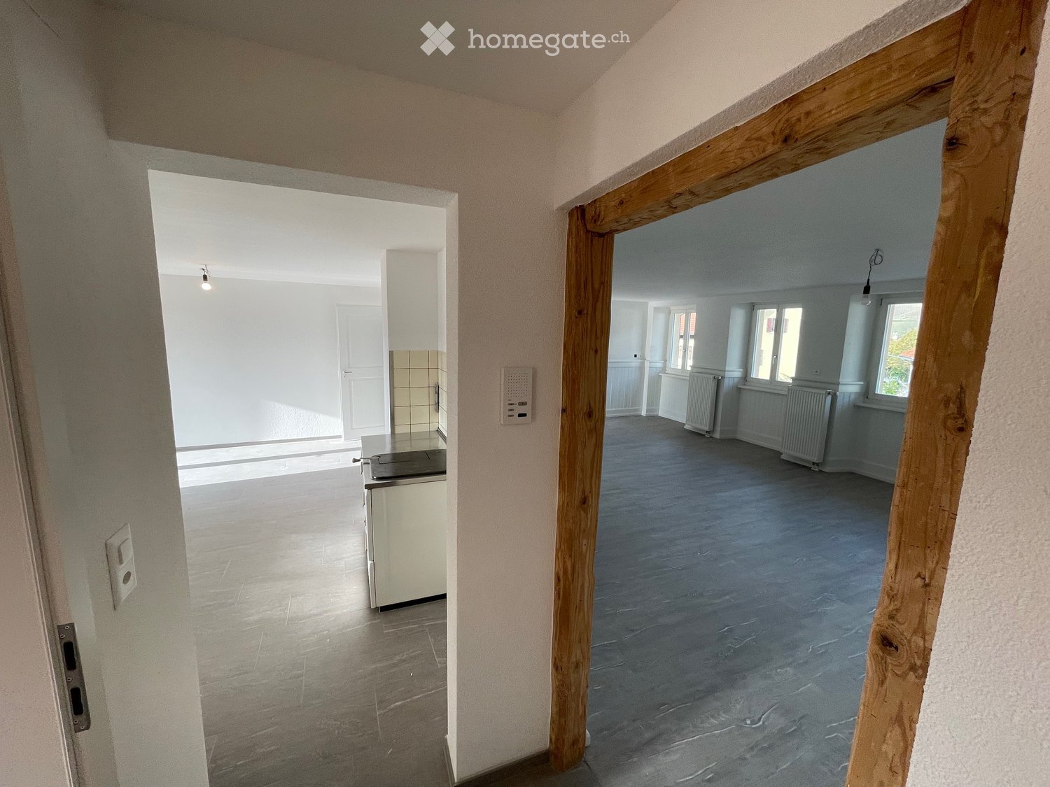 Empty room, wooden door frame, white walls, gray floor, electrical outlet, small kitchen area, radiators, and windows.