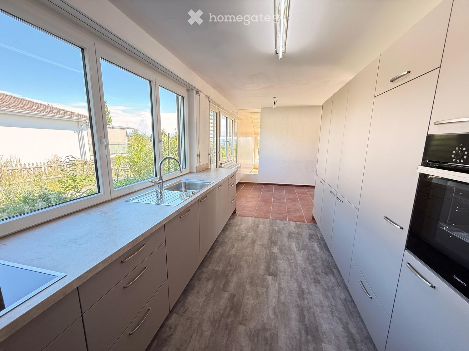 Modern kitchen with white cabinets, stainless steel sink, and built-in oven. Large windows provide natural light and a view of the garden.
