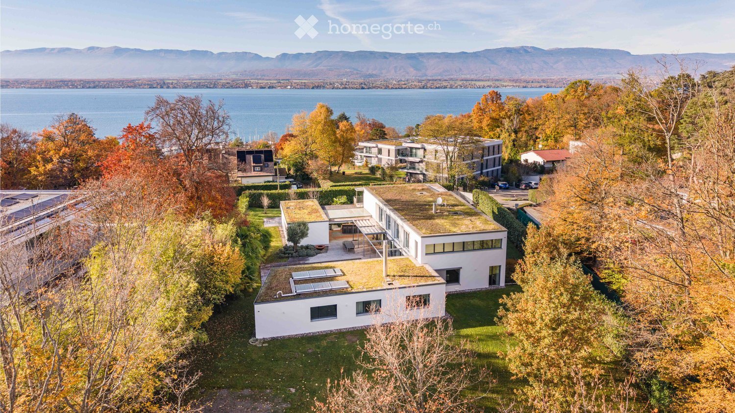 Aerial view of a modern white house with a green roof, surrounded by trees and overlooking a lake and mountains.