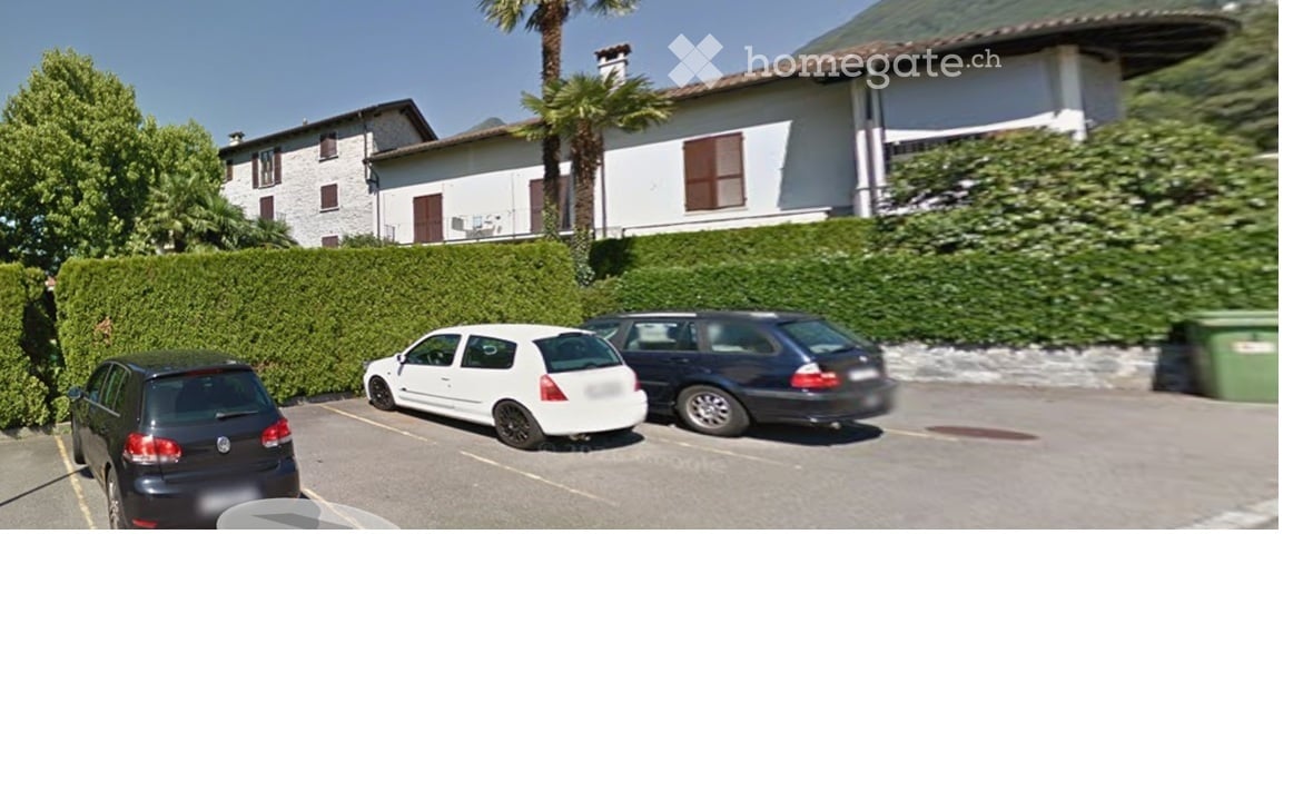 A residential building with a parking area in front, featuring several parked cars and a well-maintained hedge lining the driveway.