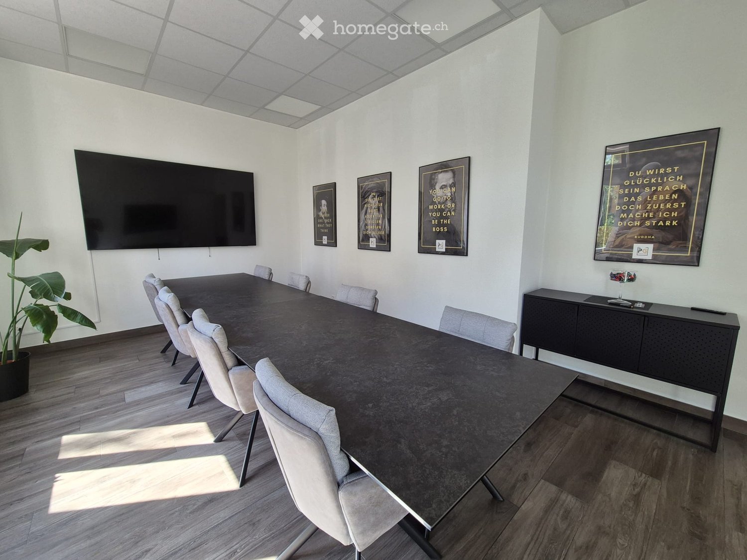 Modern conference room with a long table, chairs, a large screen, and a cabinet with a glass display. The room features a wooden floor and large windows allowing natural light.