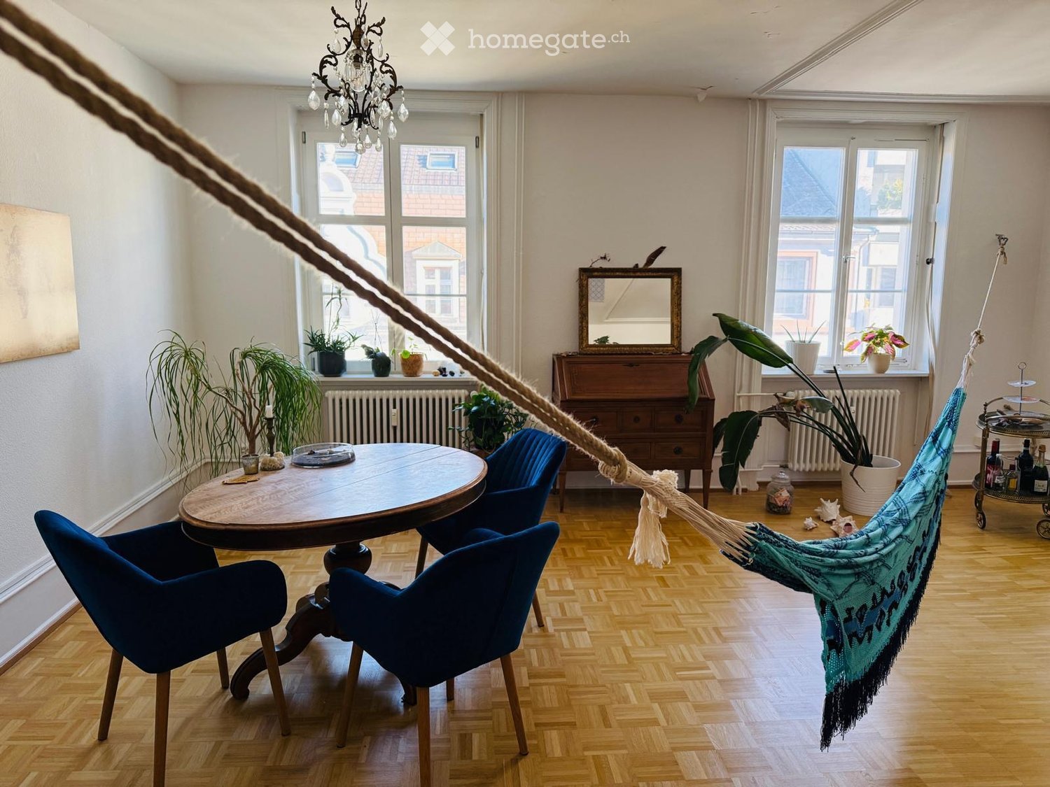 A cozy living room with wooden flooring, large windows, a round wooden table with chairs, a hanging hamaca, a wooden cabinet, and several potted plants.