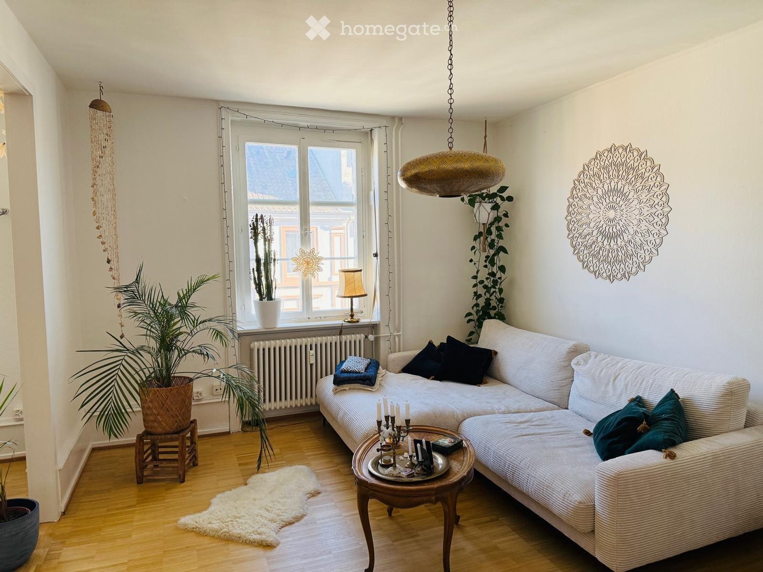 Cozy living room with a white sofa, coffee table, table lamp, and decorative items. Features wooden flooring, large window with white curtains, and several potted plants.