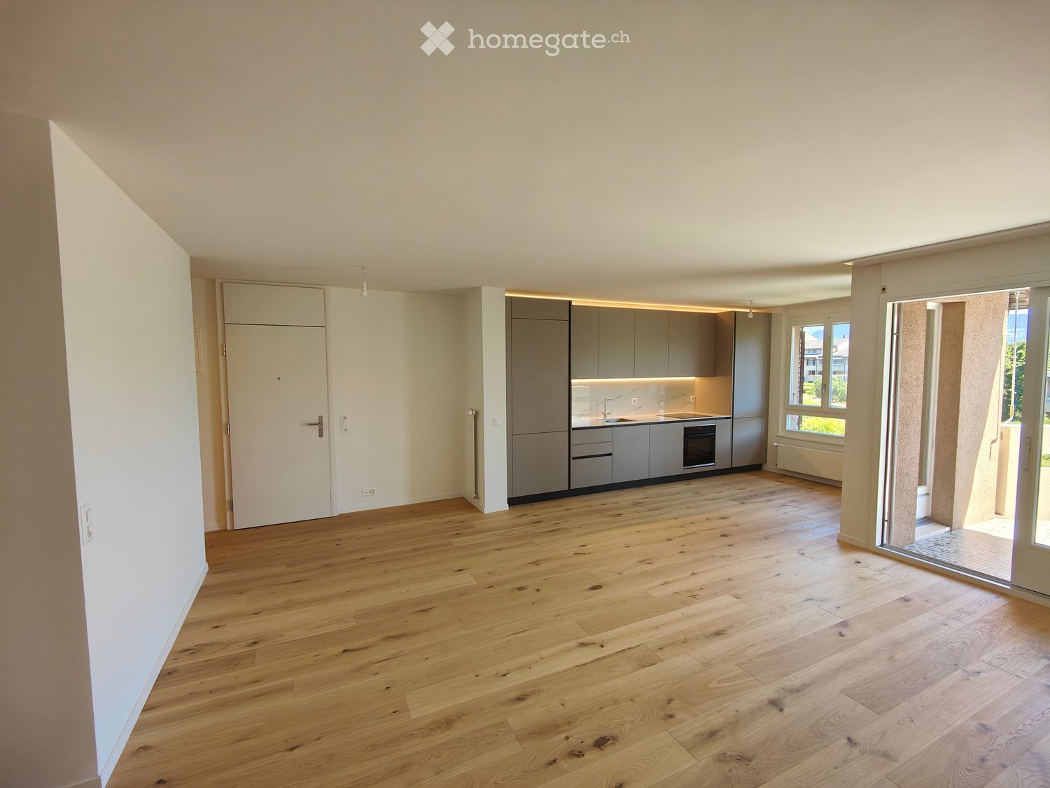 Empty room with light wooden flooring, white walls, grey kitchen cabinets, and a large window with a view of greenery.
