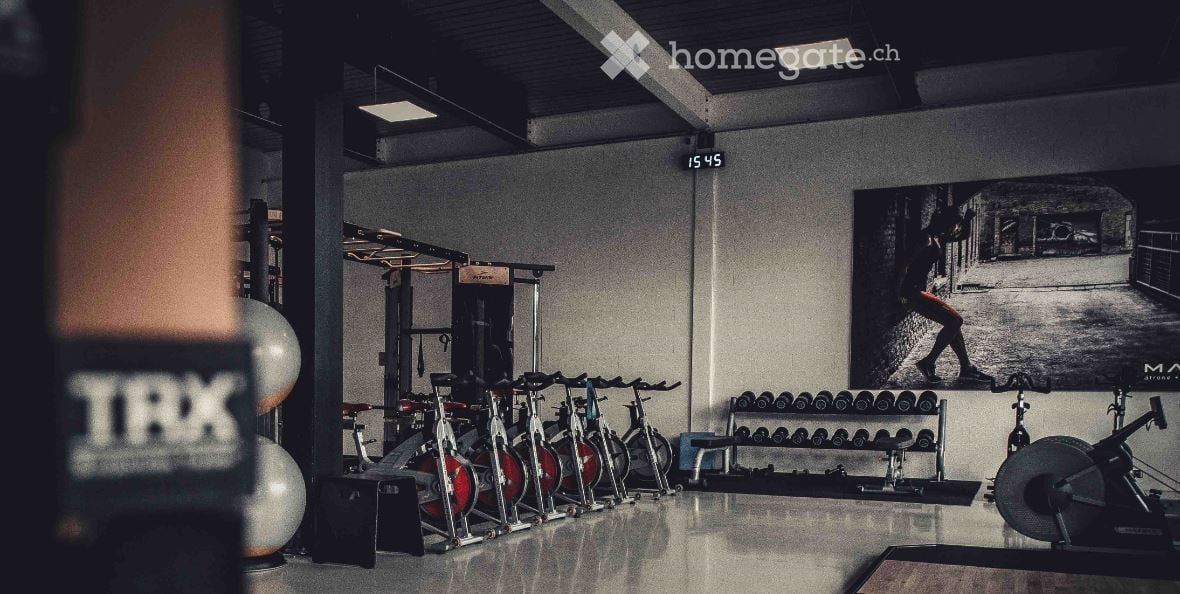 Modern gym space with multiple exercise bikes, dumbbell rack, fitness ball, and weight machine. Large black-and-white poster on wall.