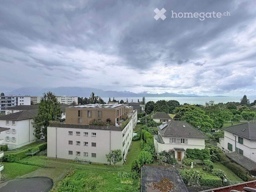 A multi-story residential building with a flat roof and modern design, surrounded by greenery and other buildings, with a scenic view of a lake and mountains in the background.