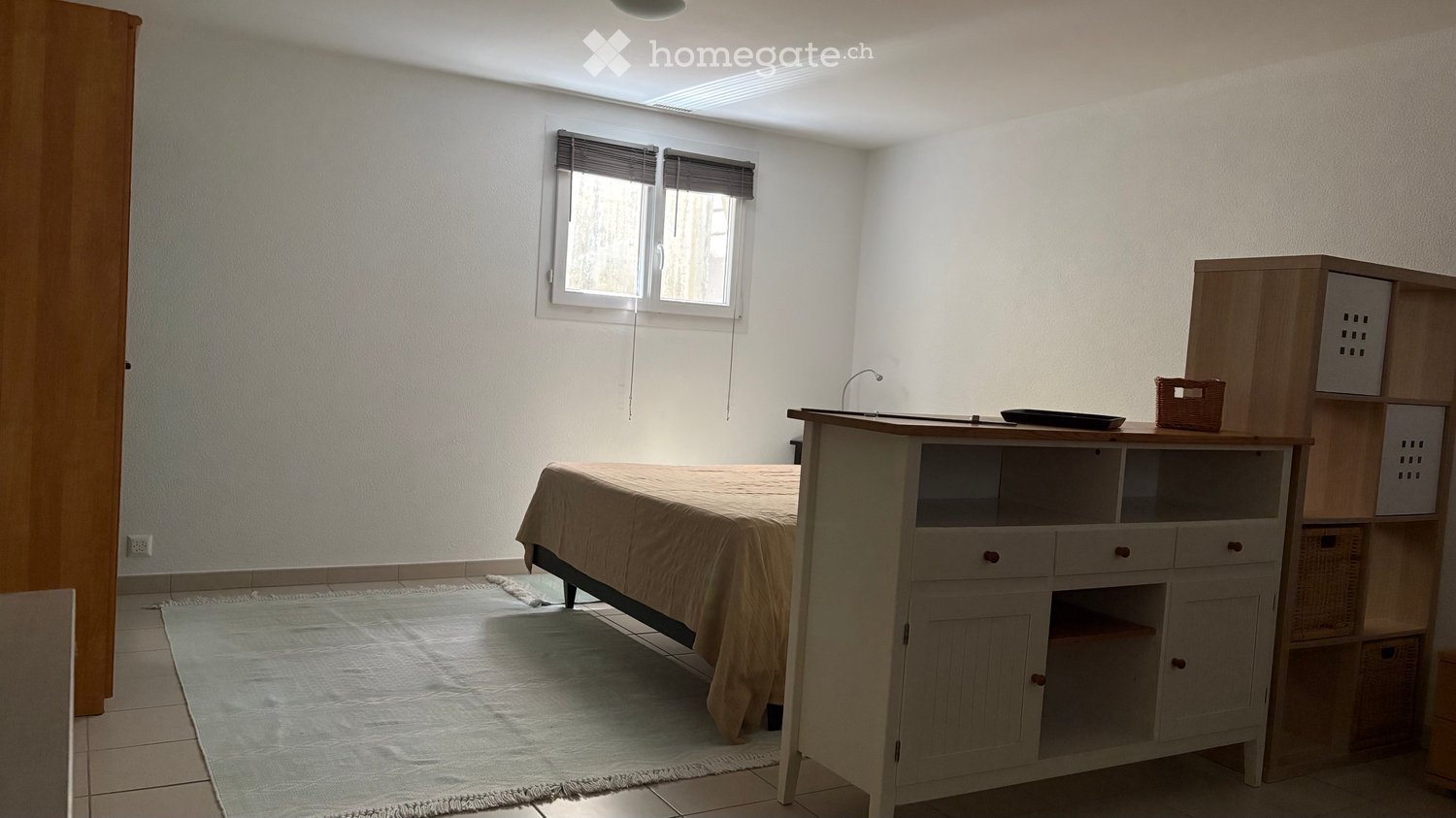 A small, furnished room with a bed, a desk, and a cabinet. The room has white walls, tiled flooring, and a window with blinds.