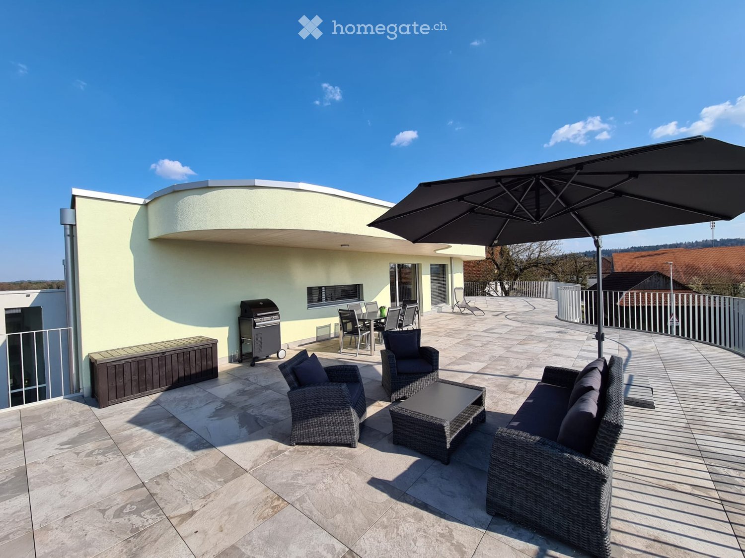 A modern outdoor patio with a large umbrella, wicker furniture including a sofa, coffee table, and chairs, a dining table with chairs, and a grill, all set on a tiled floor with a view of the surrounding landscape.