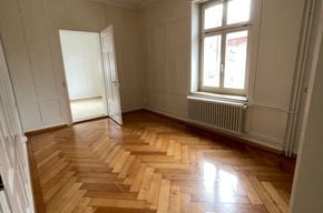 Empty room with wooden parquet flooring, white walls, a single window with radiator below, and an open door leading to another space.