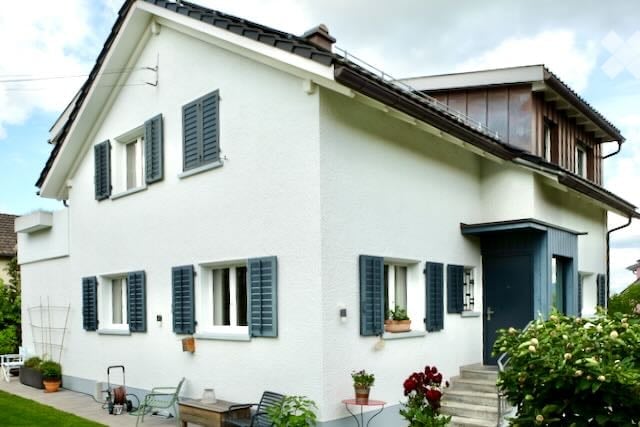 Two-story white house with blue shutters, multiple windows, and a small garden area with outdoor furniture.