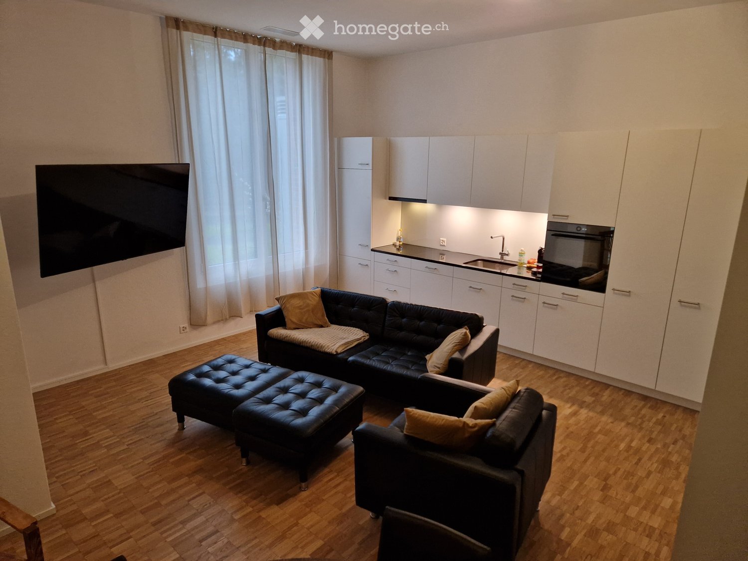Modern living room with a black leather sofa, ottoman, armchair, and a flat-screen TV. The room features a kitchen area with white cabinets, a sink, and an oven. The space is well-lit with large windows and wooden flooring.