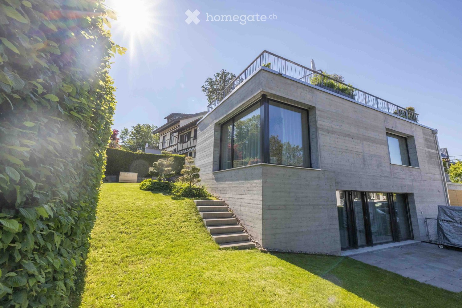 Modern two-story house with concrete facade, large windows, and a rooftop terrace. The house is surrounded by a well-maintained lawn and garden, with steps leading up to the entrance.